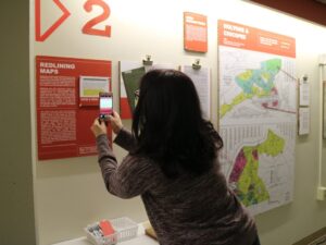 Person with black hair holds a cell phone to take a picture of a sign that says "Redlining Maps," part of the Undesign the Redline exhibit. 